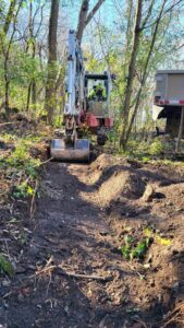 An excavator digging a trench in a wooded area for a landscaping project by Wildflower Landscapers LLC in Larsen, WI