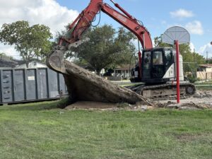 An excavator from White Star Services LLC lifting a large concrete slab with a roll-off dumpster in the background in Corpus Christi, TX.