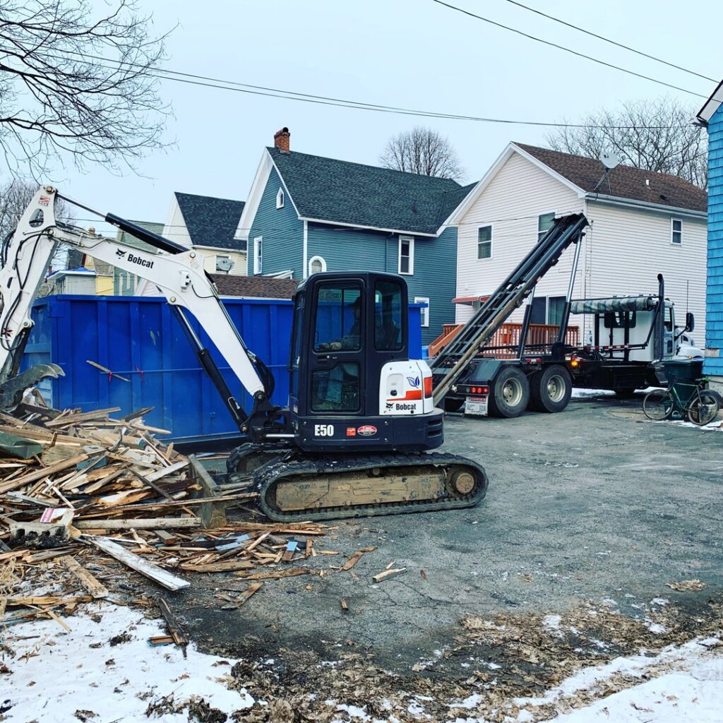 An excavator loading construction debris into an Expert Dumpster roll-off container at a job site in Rochester, NY.
