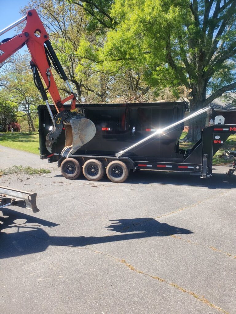 An excavator loading various debris into a black dumpster on an asphalt driveway for Standard Dumpster LLC in Rock Hill, SC.