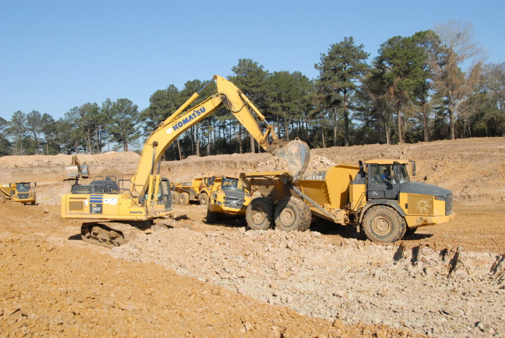 An excavator loading a dump truck with earth on a large earthmoving site, a project by Sendero Industries in Houston, TX.