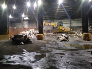 A yellow excavator actively working on a large junk pile inside the processing facility for Lemcor in Newark, NJ.