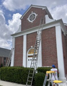 A worker on a ladder painting white trim on a brick building, a handyman painting service by Randy Overacre Painting Company in Virginia Beach, VA.