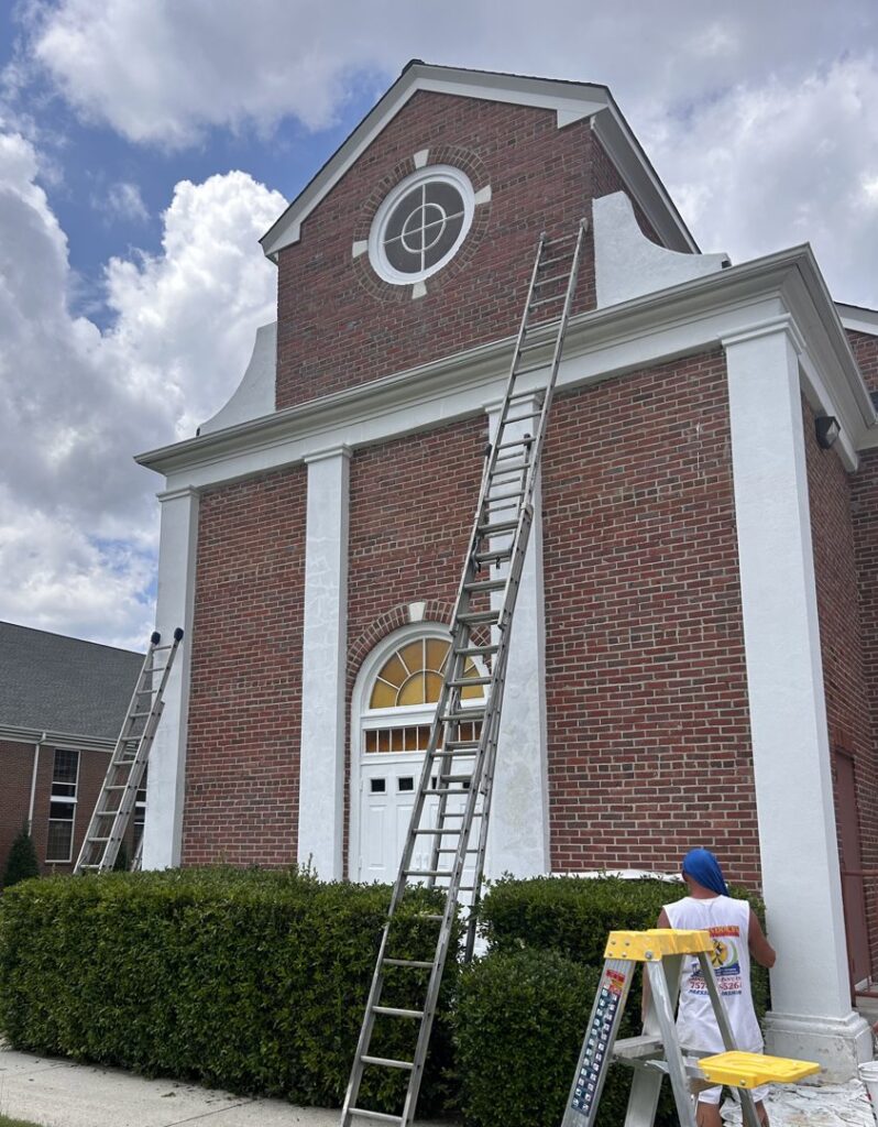 A worker on a ladder painting white trim on a brick building, a handyman painting service by Randy Overacre Painting Company in Virginia Beach, VA.