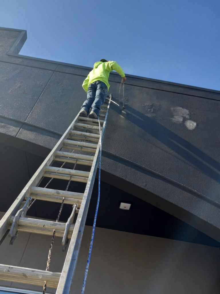 A worker performing exterior building maintenance on a tall ladder for Husky drywall LLC in Kansas City, MO.