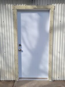 A newly installed white exterior door with a simple frame on a corrugated metal wall by My Quality Handyman in Fresno, CA.