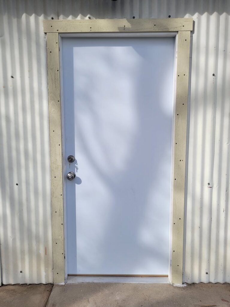 A newly installed white exterior door with a simple frame on a corrugated metal wall by My Quality Handyman in Fresno, CA.