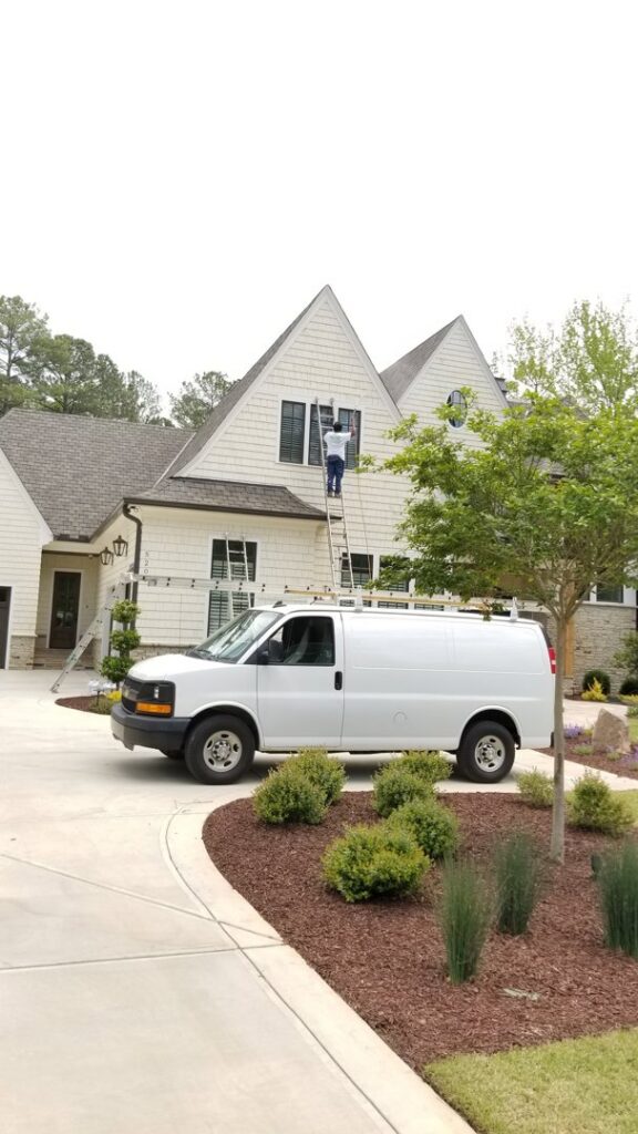 A worker on a ladder painting the exterior of a house, with the Albis Morales Painting LLC van parked nearby in Raleigh, NC.