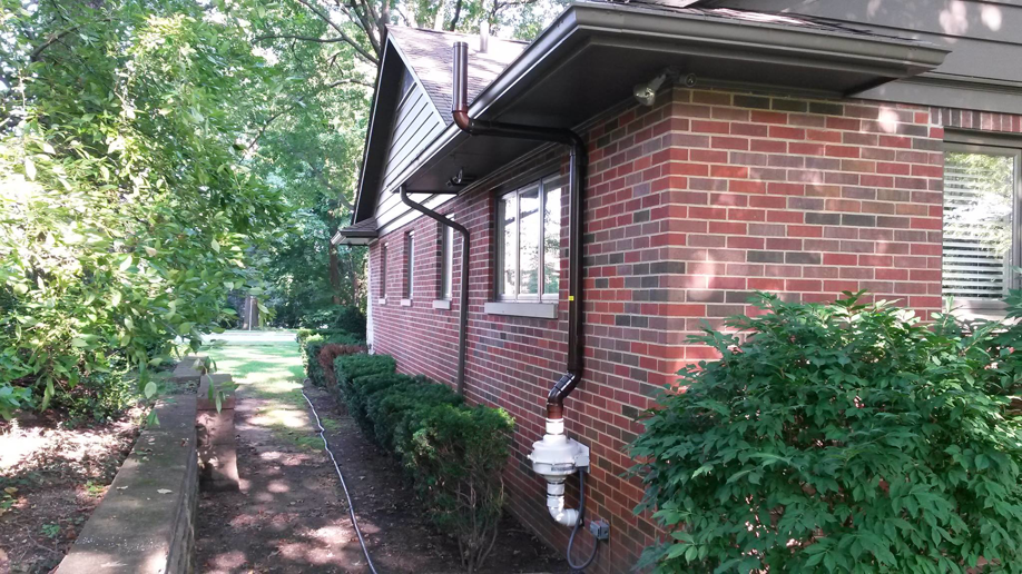An exterior dark brown radon mitigation system installed on a brick house by All Ohio Radon Services in Akron, OH.