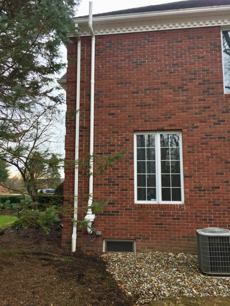 A white radon mitigation system installed on the side of a brick house by All Ohio Radon Services in Akron, OH.