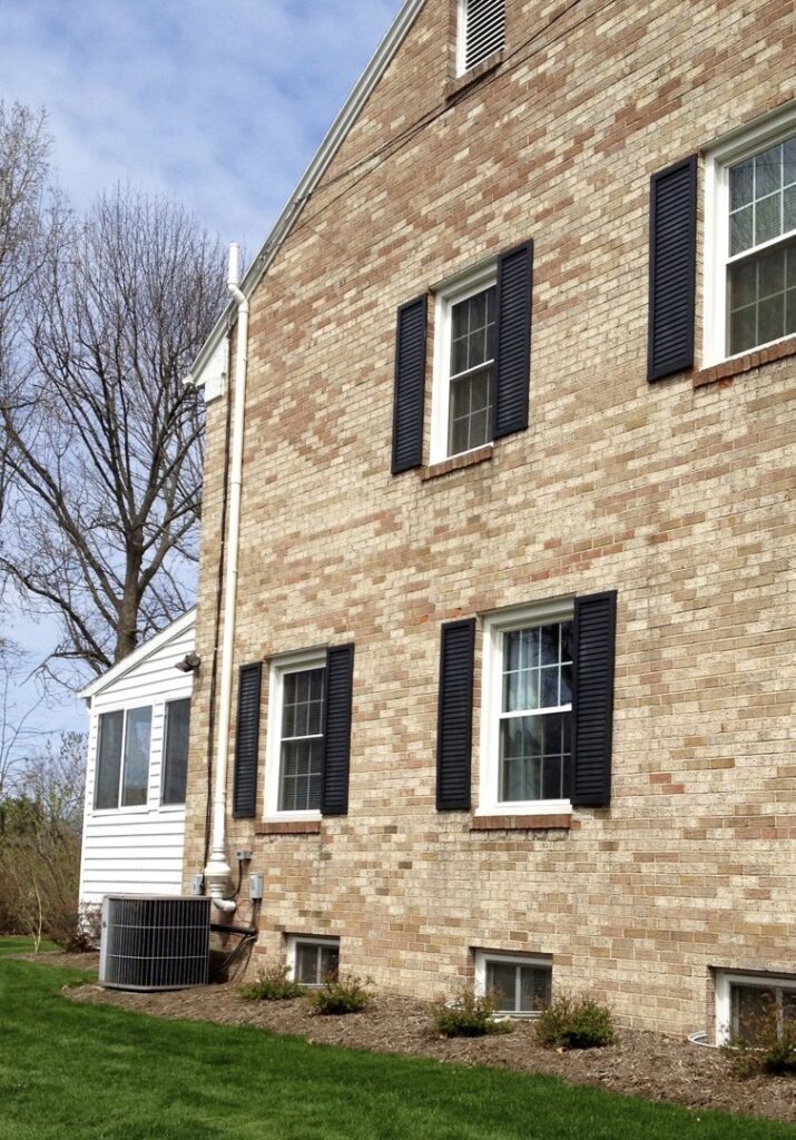 A white radon mitigation system installed on the exterior of a brick house with black shutters by All Ohio Radon Services in Akron, OH.