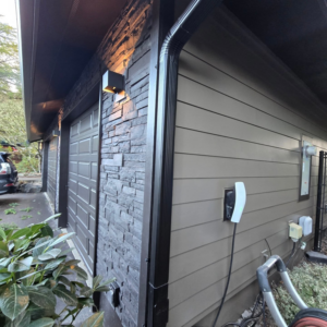 Close-up of house exterior with stone facade, new siding, downspout, and EV charger by Final Touch Construction, LLC in Tukwila, WA.