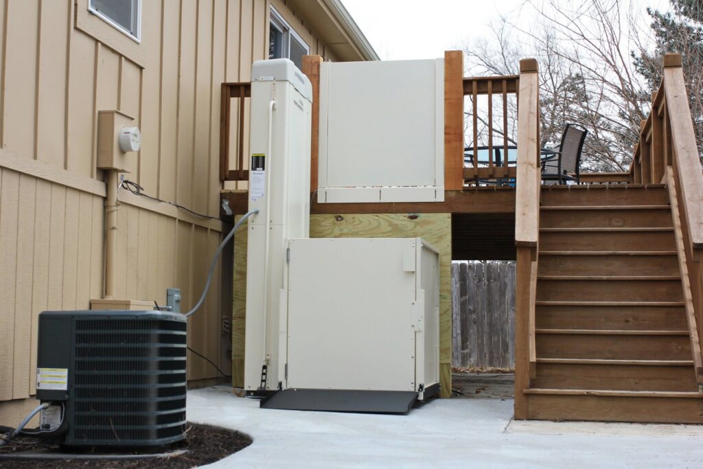 An exterior vertical platform lift installed next to a wooden deck by Thrive Homes, LLC in Olathe, KS.