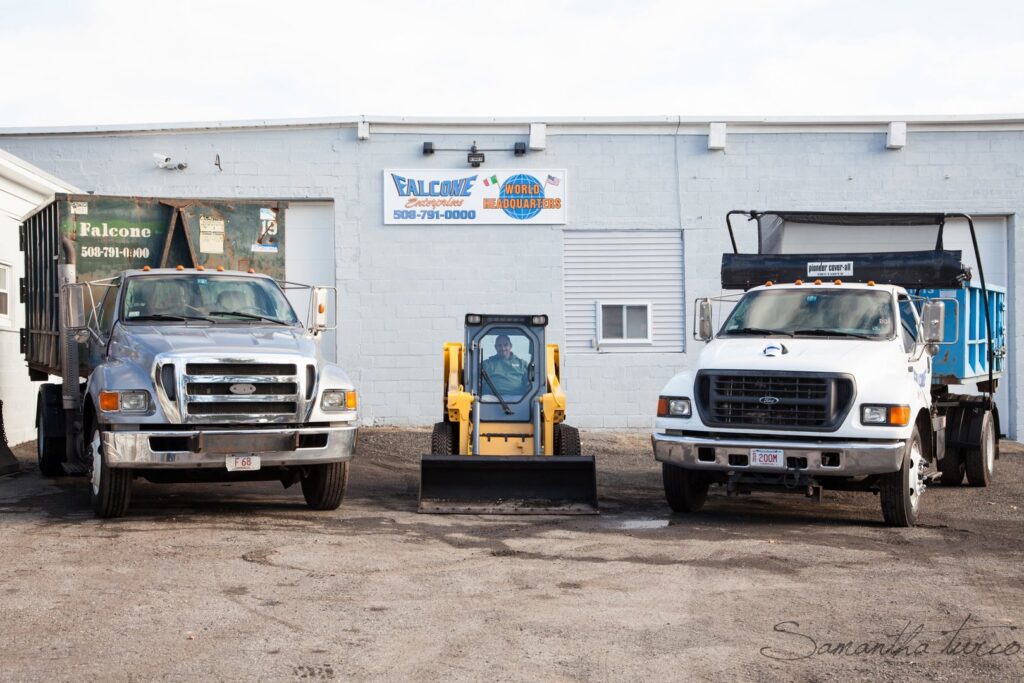 Two Falcone Enterprises junk removal trucks and a skid steer parked outside their facility in Worcester, MA.