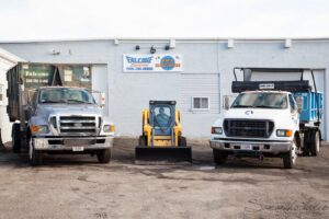 Two Falcone Enterprises junk removal trucks and a skid steer parked outside their facility in Worcester, MA.