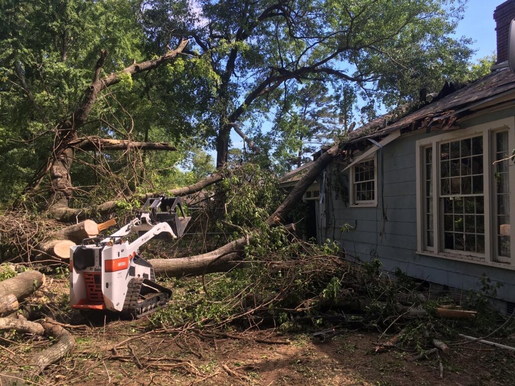 A large tree fallen onto a house, with a Bobcat skid steer clearing debris during storm cleanup by Tri-County Tree And Restoration in Jackson, MS.