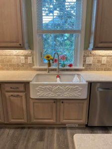 A newly installed white farmhouse kitchen sink with stone backsplash and light cabinets by Nailed It Custom Remodeling in Chesapeake, VA.
