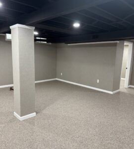 A finished basement room with new carpeting, a wrapped column, and a dark ceiling by Impressive Basements in Westland, MI.