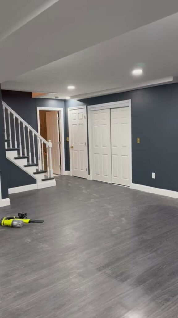A newly finished basement room with dark gray walls, new flooring, and a white staircase, completed by Men Construction in Lowell, MA.