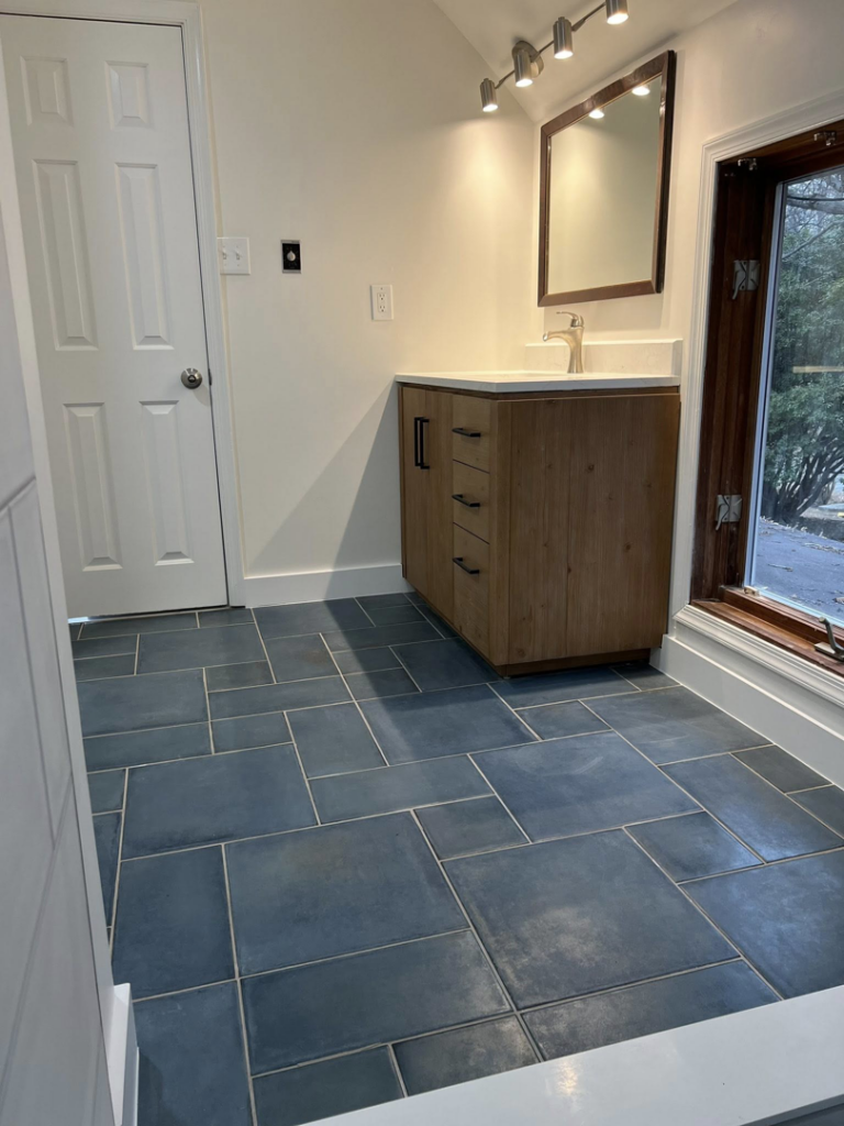 A finished bathroom renovation showcasing a modern wooden vanity, mirror, and dark blue floor tiles by Great Home Construction LLC in Annapolis, MD.