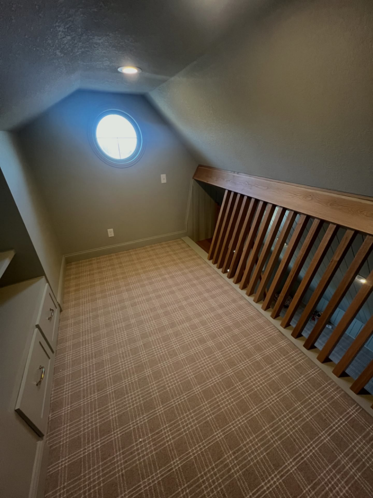 A newly finished loft space with plaid carpet, an oval window, and built-in storage by H4 Construction and Remodeling in Blue Springs, MO.