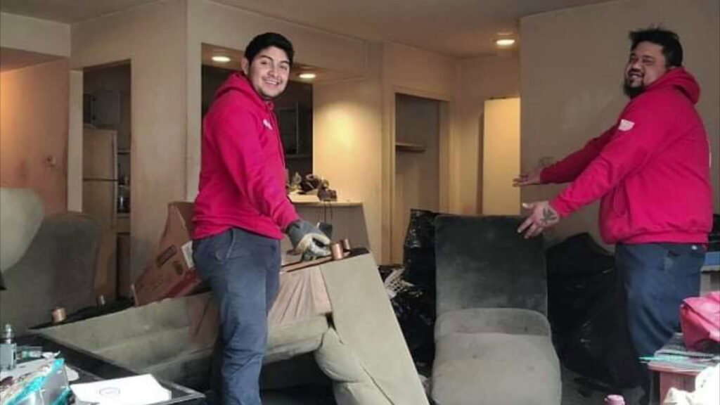 Two Fire Dawgs Junk Removal employees stand in a cluttered room, ready to begin a junk removal job in Bloomington, IN.