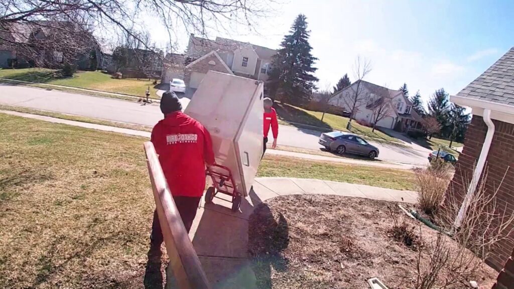 Fire Dawgs Junk Removal employees carefully move a large appliance on a dolly down a driveway in Bloomington, IN.