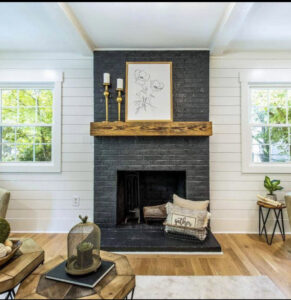 A remodeled living room featuring a black brick fireplace with a rustic wood mantel and shiplap walls by Pro Finishes NC, LLC in Raleigh, NC.