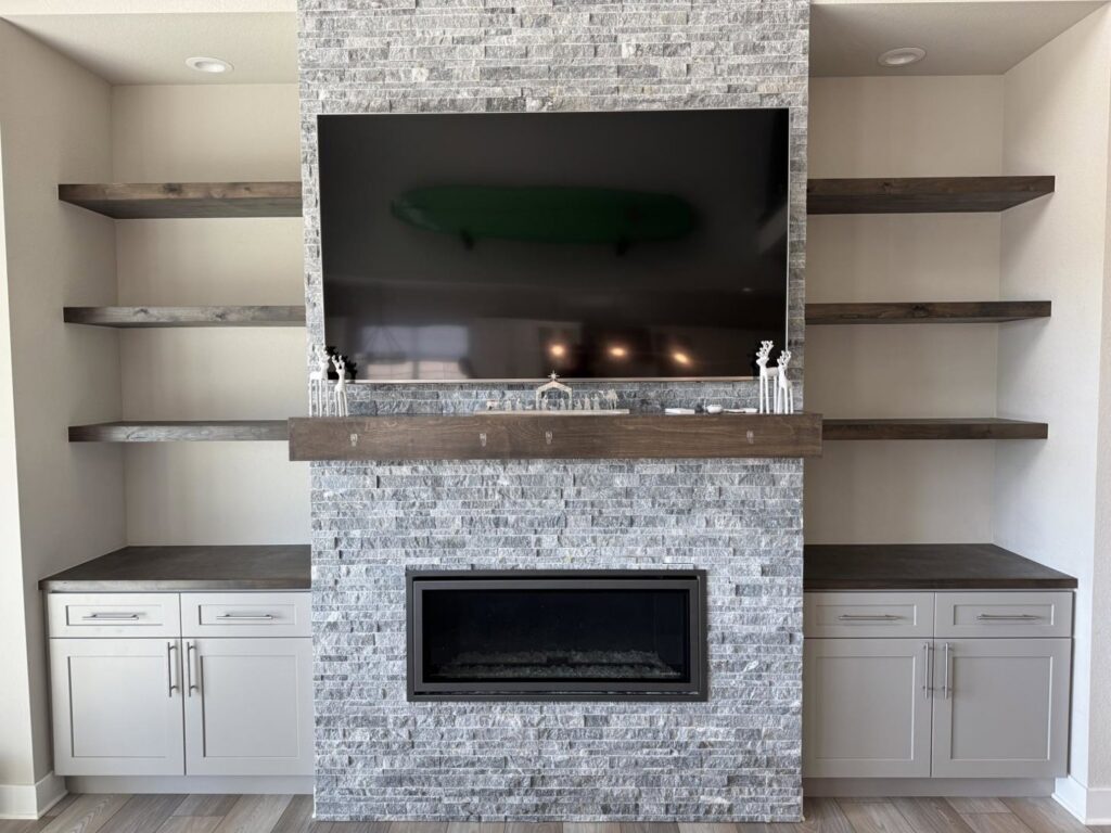 A living room with a stone-faced fireplace, mounted TV, floating shelves, and lower cabinets installed by Colorado ProBuilt in Aurora, CO