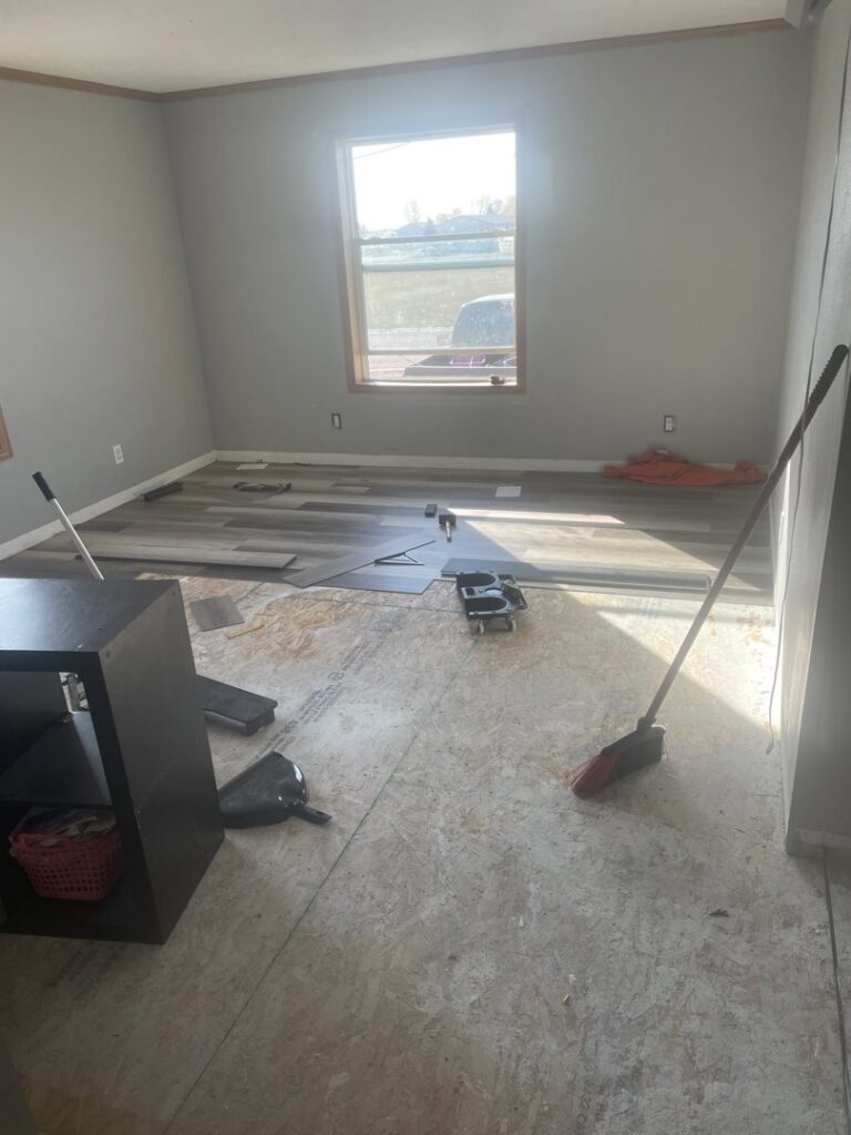 A room with subfloor exposed and new flooring planks laid out, showing a flooring installation in progress by Knutson's Property Maintenance Llc in Williston, ND.
