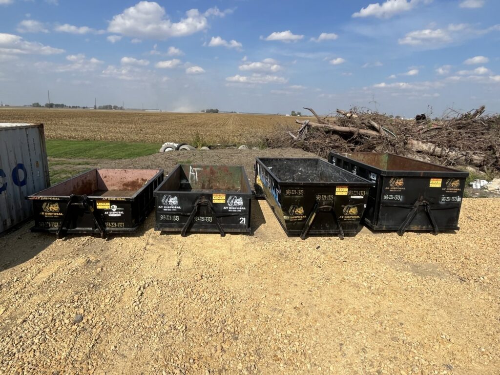 Four small roll-off dumpsters from AT Disposal lined up in a rural setting in Dewitt, IA.