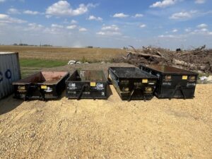 Four small roll-off dumpsters from AT Disposal lined up in a rural setting in Dewitt, IA.