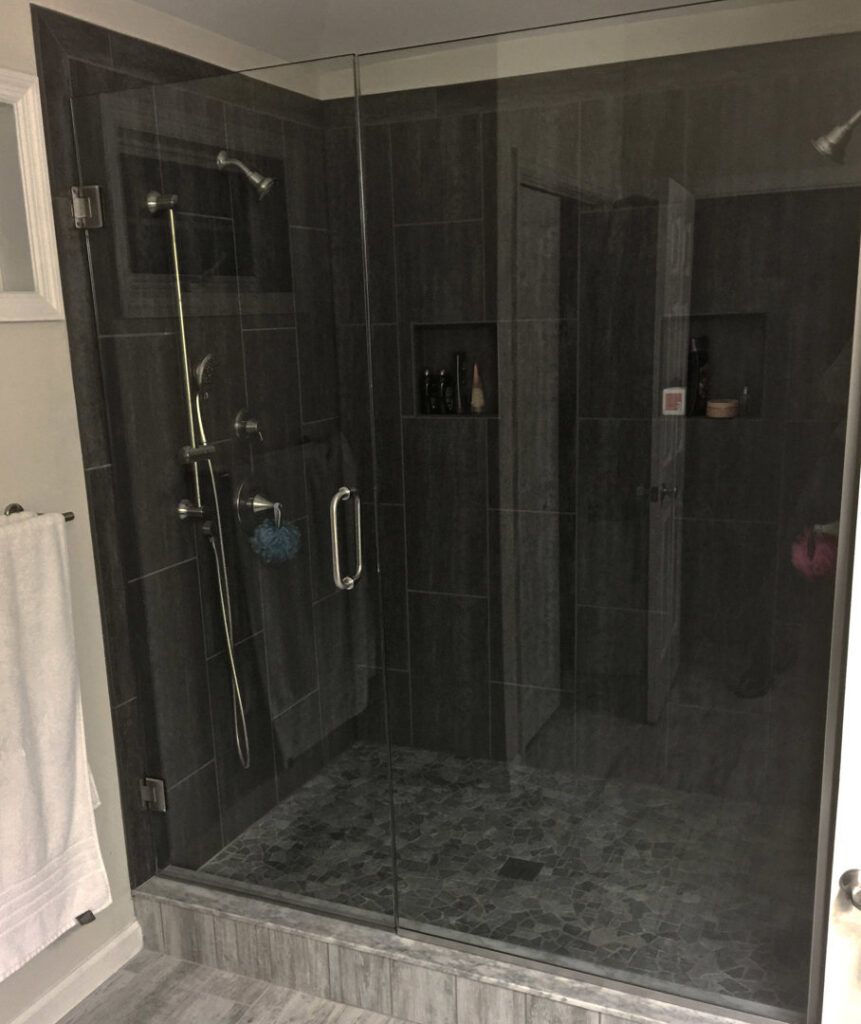 A frameless glass shower with dark tiled walls and a pebble floor, expertly installed by Lehigh Shower Glass in Bethlehem, PA