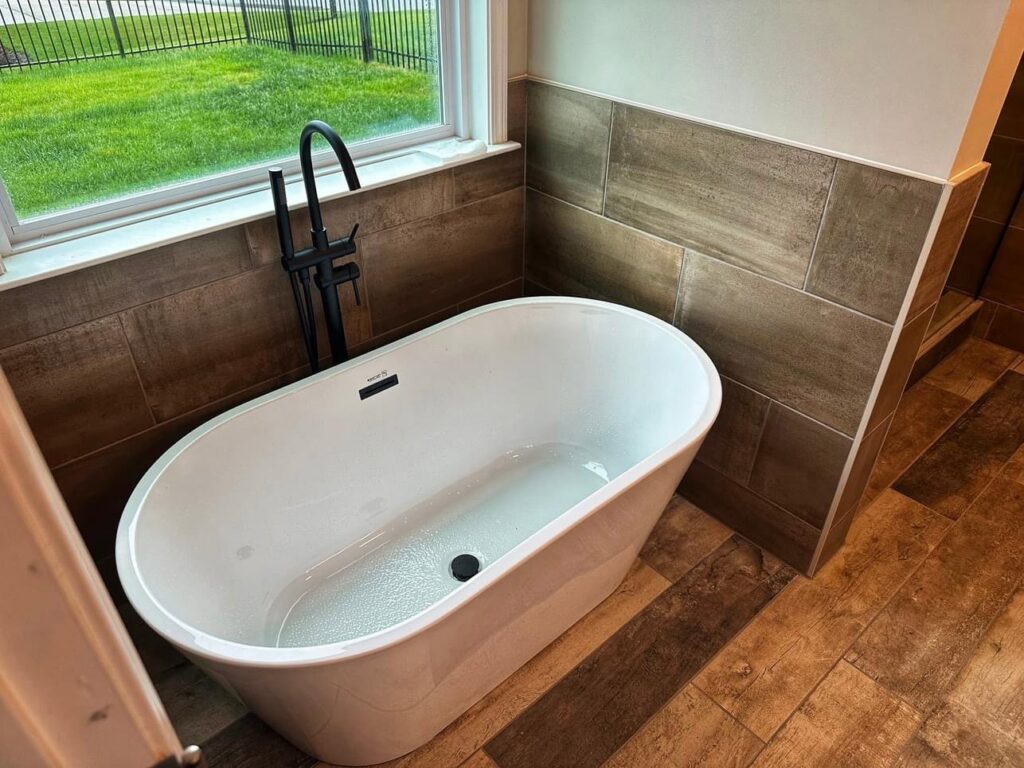 Freestanding bathtub with a black floor-mounted faucet, surrounded by wood-look tiles, installed by Supreme Tile & Flooring in Charlotte, NC.
