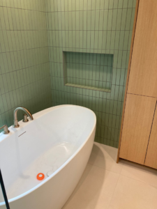 A sleek freestanding bathtub positioned next to a wall with green subway tiles and a built-in niche by Tess Tile By Design in Kansas City, MO.