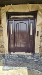 A residential front door being professionally painted brown, with surrounding areas taped off by Acosta Painting LLC in Jonesboro, AR.