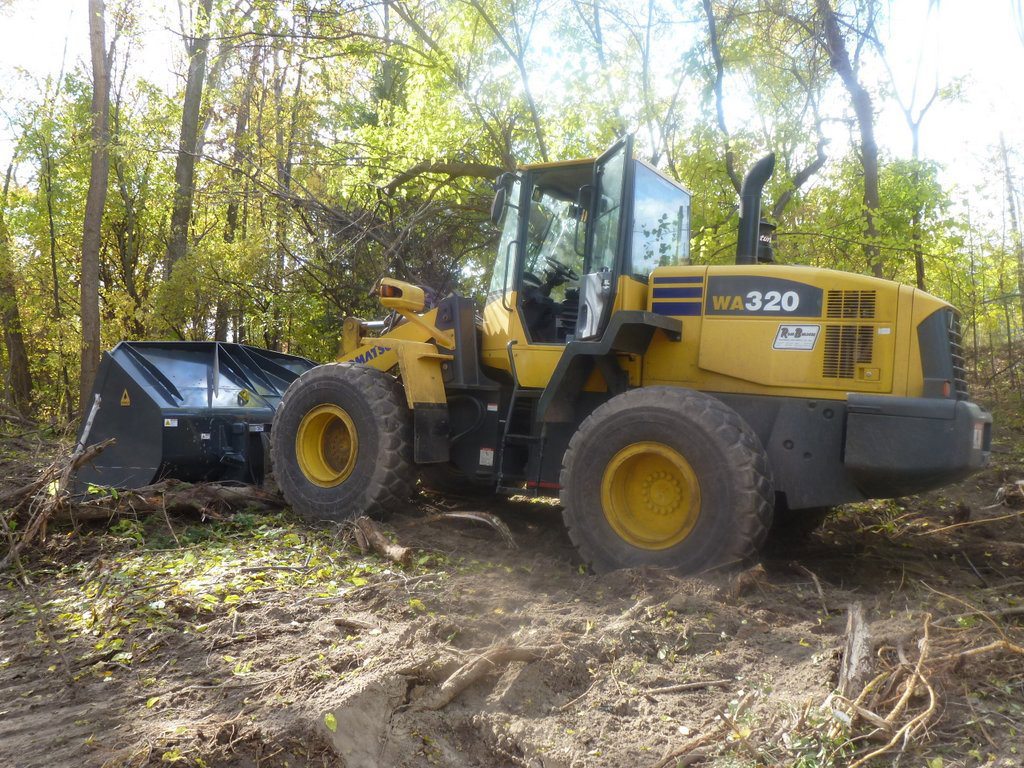 A large yellow front-end loader with a mulching attachment clearing brush and small trees in a wooded area for Hofeling Enterprises in Lincoln, NE.