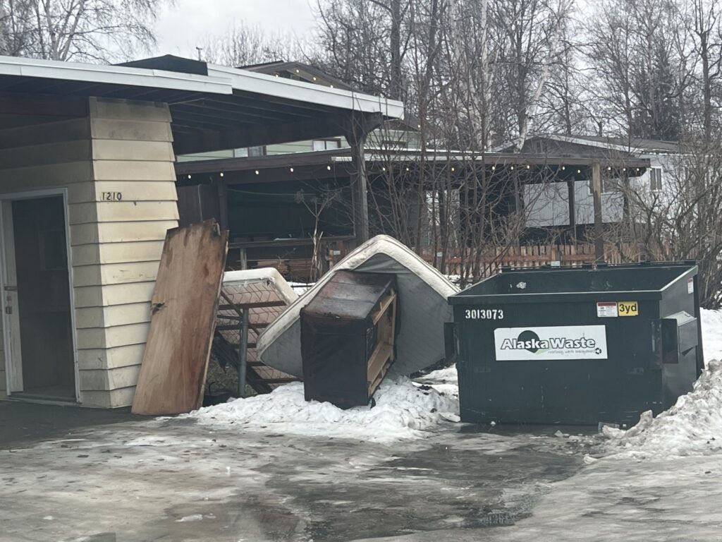 A pile of old furniture, including a mattress and dresser, and other junk next to a dumpster by Alaska Waste in Anchorage, AK.