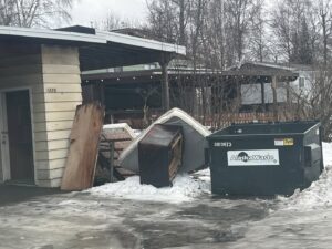 A pile of old furniture, including a mattress and dresser, and other junk next to a dumpster by Alaska Waste in Anchorage, AK.