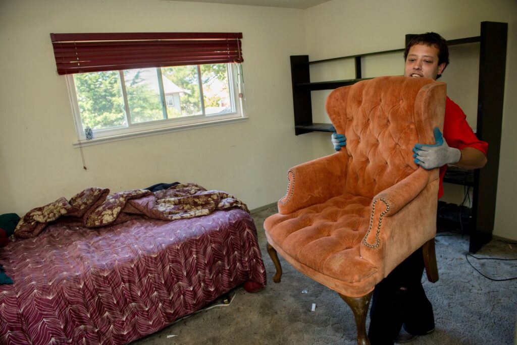 A Junk King Plano, TX employee carefully carrying a large upholstered armchair out of a room for removal.