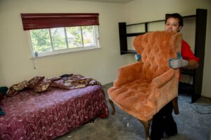 A Junk King Plano, TX employee carefully carrying a large upholstered armchair out of a room for removal.