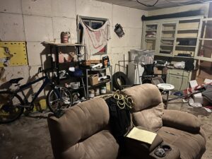 A cluttered garage or basement room with a bicycle, recliner, and shelves, requiring junk removal by Feelin Good Junk Removal & Estate Sales at 1111 Debra Lane.