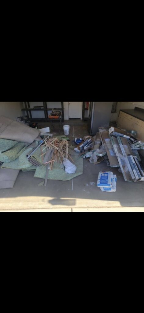 A garage floor covered with construction debris, old carpet, wood scraps, and various junk items for removal by The Junkporter Junk Removal in Las Vegas, NV.