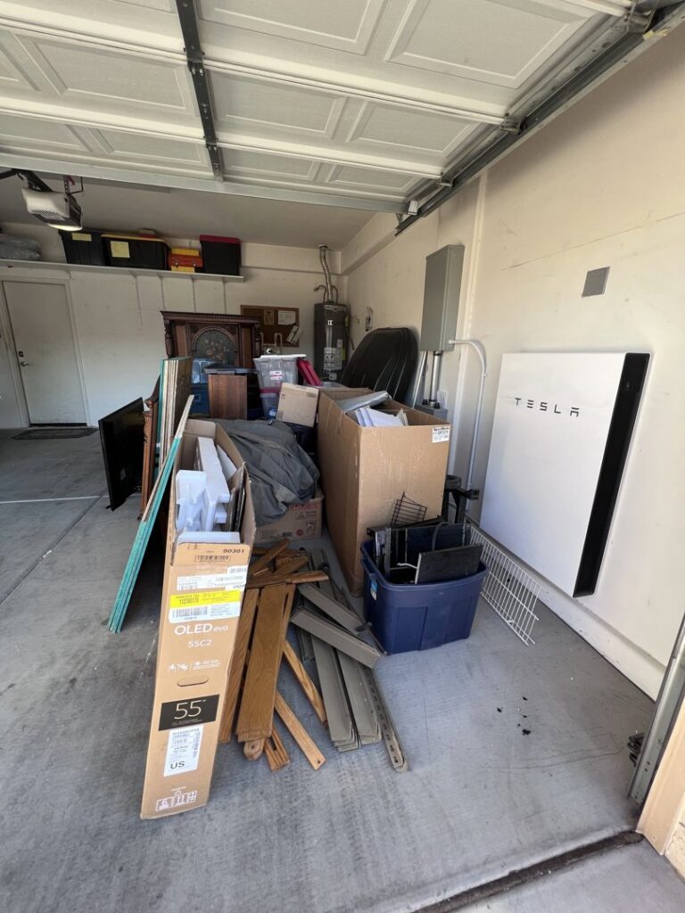A garage filled with boxes, furniture, wood scraps, and a Tesla Powerwall unit, ready for junk removal by MLG Strong Hauling & Junk Removal LLC in Henderson, NV.
