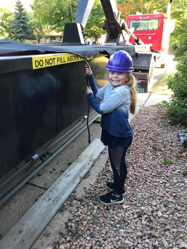 A young girl in a hard hat standing next to a large junk removal dumpster from Minnesota Waste and Transfer in Champlin, MN