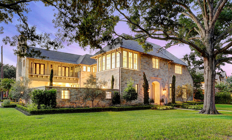 A grand stone house exterior with a spacious lawn and mature trees at dusk, constructed by Allan Edwards Builder Inc in Houston, TX.