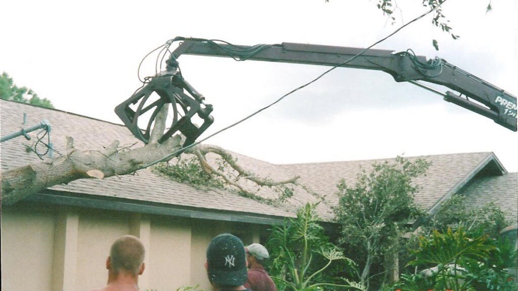 A grapple truck lifting a large tree branch over a house roof during tree service by Tim's Tree Service in Cape Coral, FL