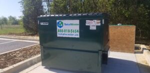 A green commercial dumpster from Total Waste placed in a parking lot surrounded by trees in Baltimore, MD.