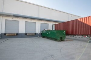 A green roll-off dumpster placed near loading docks and a shipping container, used for junk removal by Waste Master Dumpster Rental Orlando, FL.