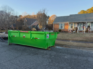 A green Maxx-D dumpster on a street with an excavator in the background, provided by Great Bridge Bins in Norfolk, VA.
